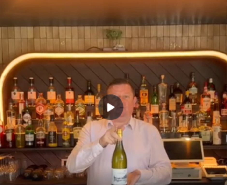 A man in a light pink shirt stands behind a bar holding a bottle of white wine, with various liquor bottles displayed on shelves in the background.