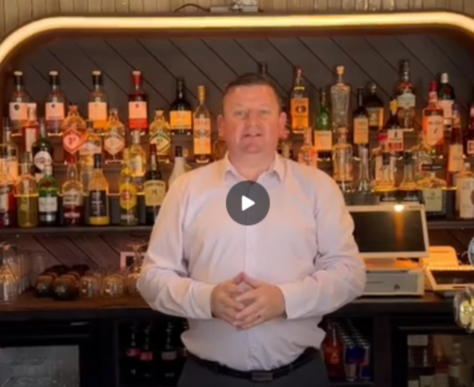 A man in a light shirt stands behind a bar with shelves of assorted liquor bottles and a cash register visible in the background.