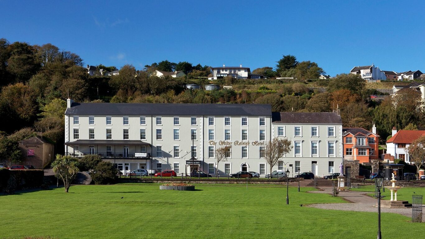 A large, white hotel building with several windows faces a green lawn under a clear blue sky, with houses and trees on the hillside behind it.
