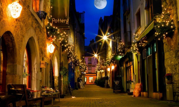 A narrow cobblestone street with festive string lights, shopfronts, and a full moon glowing in the twilight sky above.