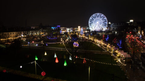A city park at night is decorated with colorful lights, illuminated cone-shaped trees, and a large lit Ferris wheel in the background.