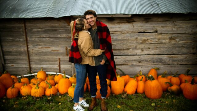 A couple stands in front of a wooden shed, surrounded by pumpkins. They are embracing and sharing a plaid blanket, with grass beneath them.