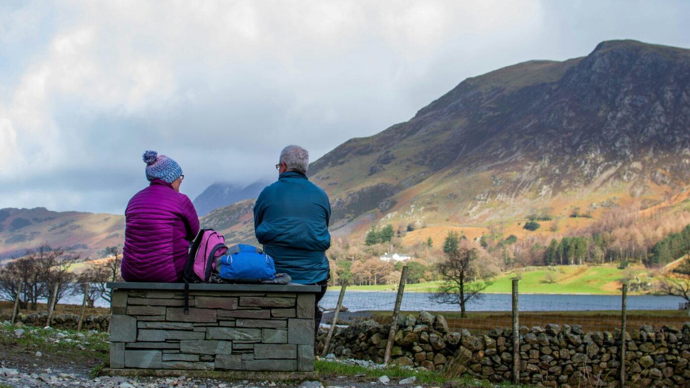 Two people in jackets sit on a stone bench, facing a scenic landscape of hills, a lake, and cloudy skies.