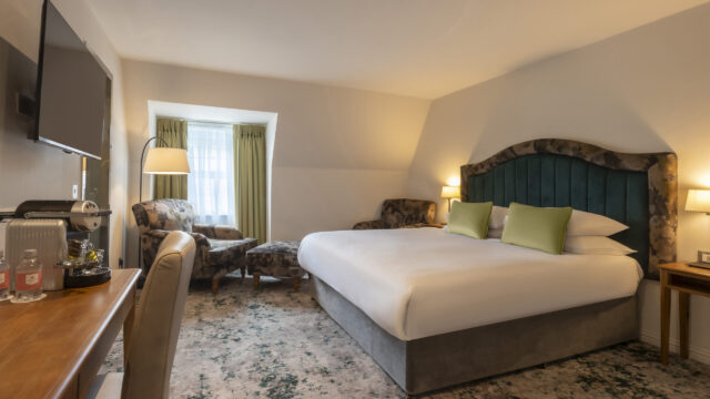 A hotel room with a large bed, green headboard, two green pillows, armchairs by a window, a desk with a chair, a TV on the wall, and lamps on either side of the bed.