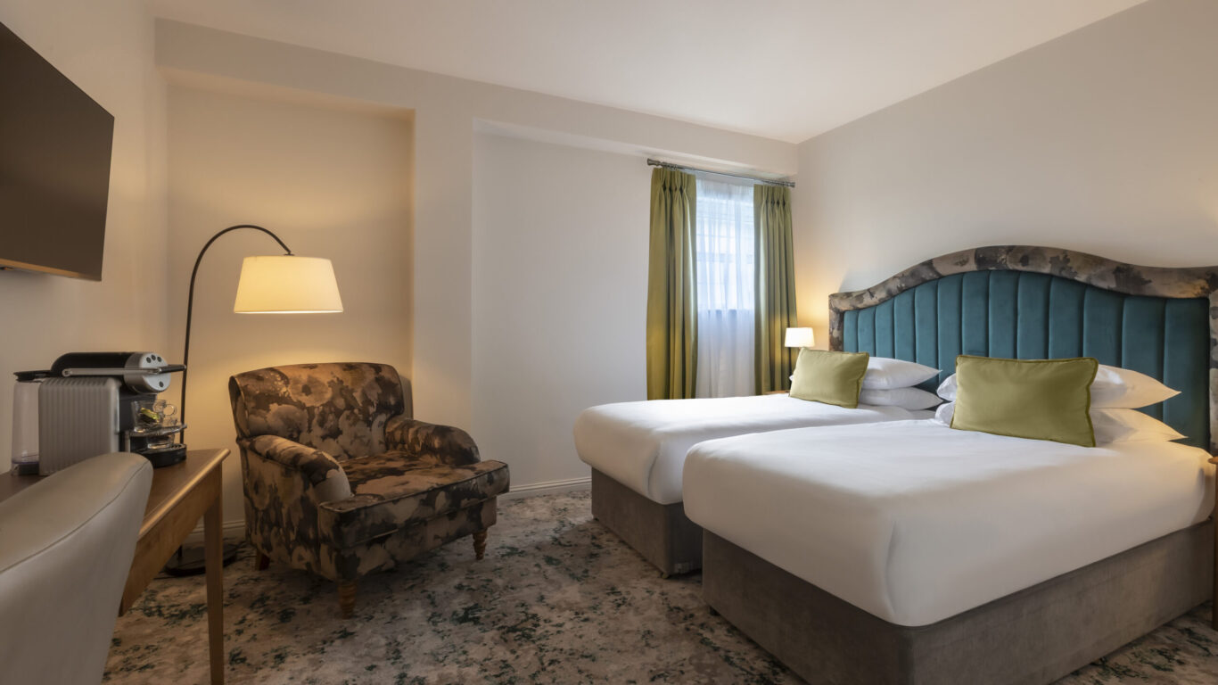 A hotel room with two twin beds, a green upholstered headboard, green curtains, an armchair, a floor lamp, a TV, and a coffee machine on a wooden table.