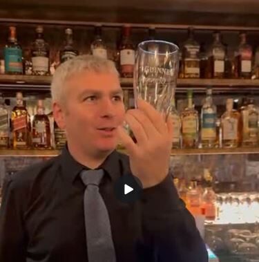A bartender wearing a black shirt and gray tie holds up an empty Guinness glass in front of a bar stocked with liquor bottles.