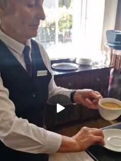 A server in a vest and tie holds a steaming cup of coffee on a saucer, preparing to place it on a tray in a dining area.