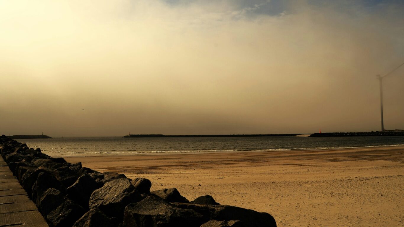 A sandy beach bordered by a rocky seawall, with calm water and a hazy sky overhead.