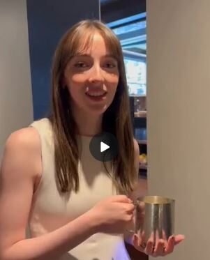 A woman with straight brown hair wearing a sleeveless white top holds a silver cup and stands indoors near a wall.