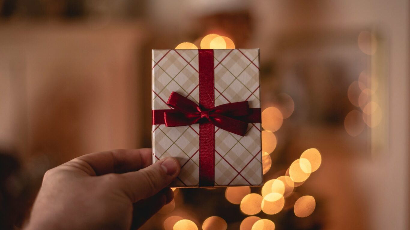 A hand holds a small, wrapped gift box with a red ribbon bow. Blurred warm lights are visible in the background.