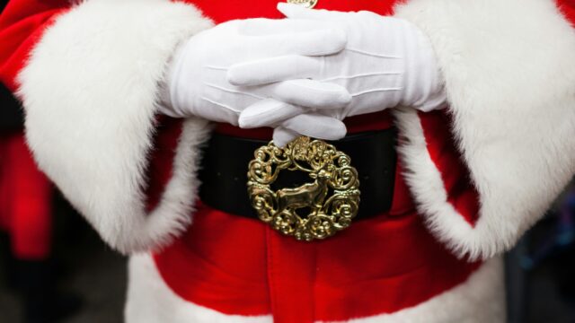Close-up of a person in a Santa Claus suit with white gloves, red coat with white fur trim, and a black belt featuring a gold reindeer buckle.