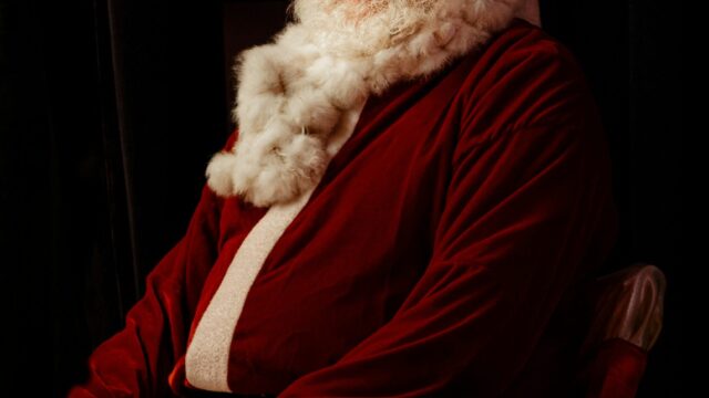 An older man dressed in a red Santa Claus costume with a white beard sits and smiles indoors.