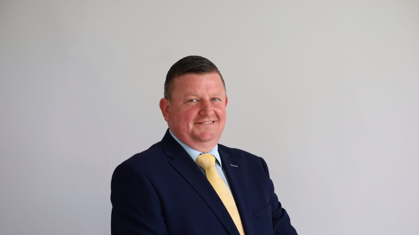 A man in a dark blue suit and yellow tie stands against a plain light-colored background, looking at the camera and smiling.